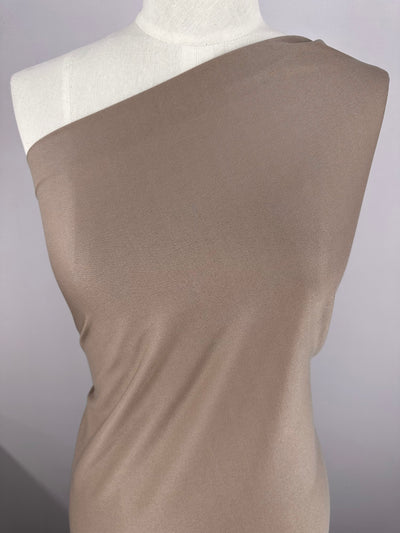 A mannequin is dressed in a one-shoulder, taupe-colored fabric draped elegantly. The lightweight fabric has a smooth texture and is made of 100% polyester, covering the mannequin diagonally and leaving one shoulder exposed. The background features Interlock - Ash - 150cm by Super Cheap Fabrics, showcasing an ash gradient from light to dark gray.