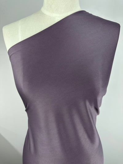 A mannequin is draped with a one-shoulder garment made of smooth, light purple 100% polyester fabric. The *Interlock - Mauve - 150cm* from **Super Cheap Fabrics** appears soft and slightly reflective, showcasing minimal wrinkles and a sleek texture. The background is neutral and simple, focusing attention on the lightweight fabric.