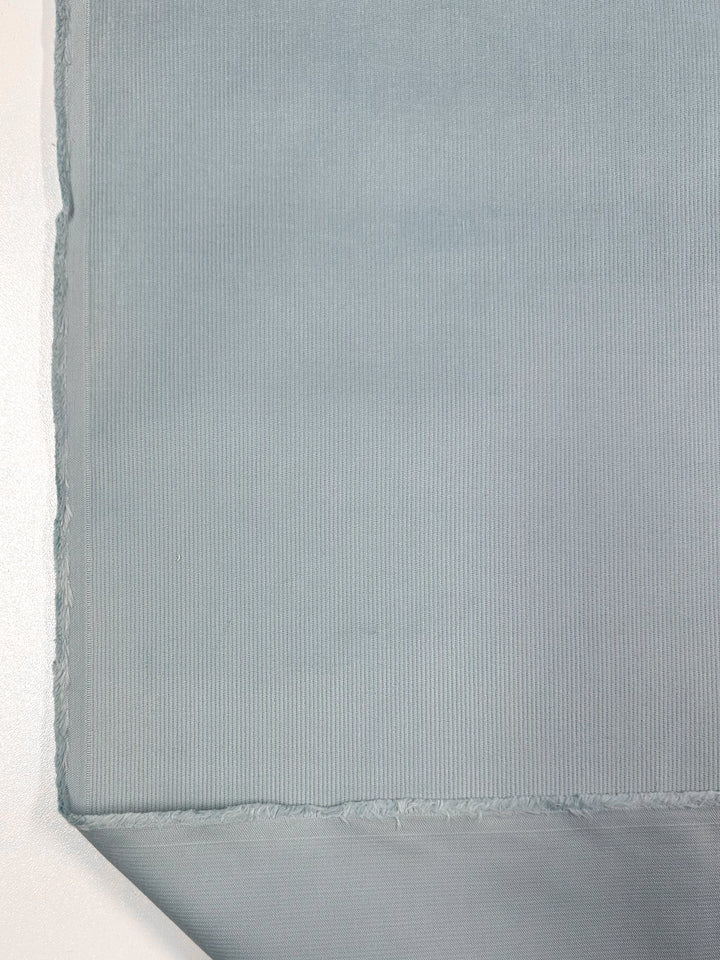 A close-up image of Spa Blue Micro Wale Corduroy fabric by Super Cheap Fabrics, showcasing its subtle vertical ribbed texture. The edge of the 145cm wide fabric is visible at the bottom left, revealing a rough, unfinished seam. The material appears soft and slightly reflective, making it perfect for winter clothing or home decor.