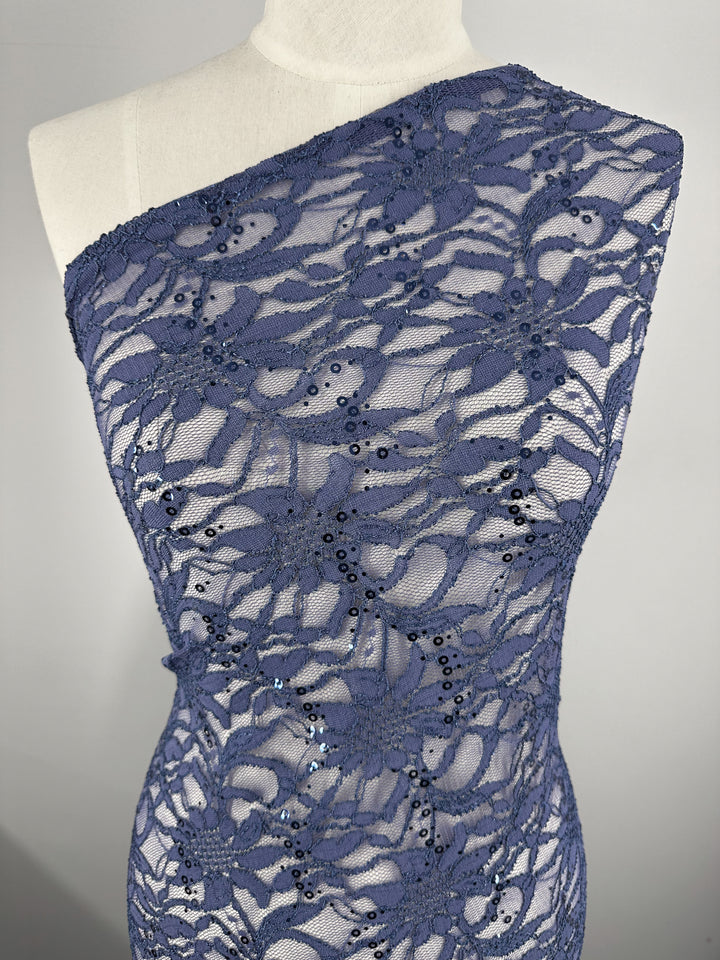 A close-up of a mannequin showcases "Evening Lace - Bengu - 135cm" by Super Cheap Fabrics, featuring a one-shoulder dress made from sheer, navy lace. The 100% polyester fabric is adorned with an elaborate floral pattern and small, subtle sequins scattered throughout, creating a textured and elegant appearance.