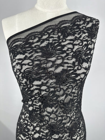 A close-up of a dress form draped in Evening Lace - Twinkle - 133cm by Super Cheap Fabrics. The black sheer fabric, adorned with floral lace patterns and occasional sequins, creates a detailed and elegant design on the lightweight polyester material. The dress form is displayed against a plain, white background.