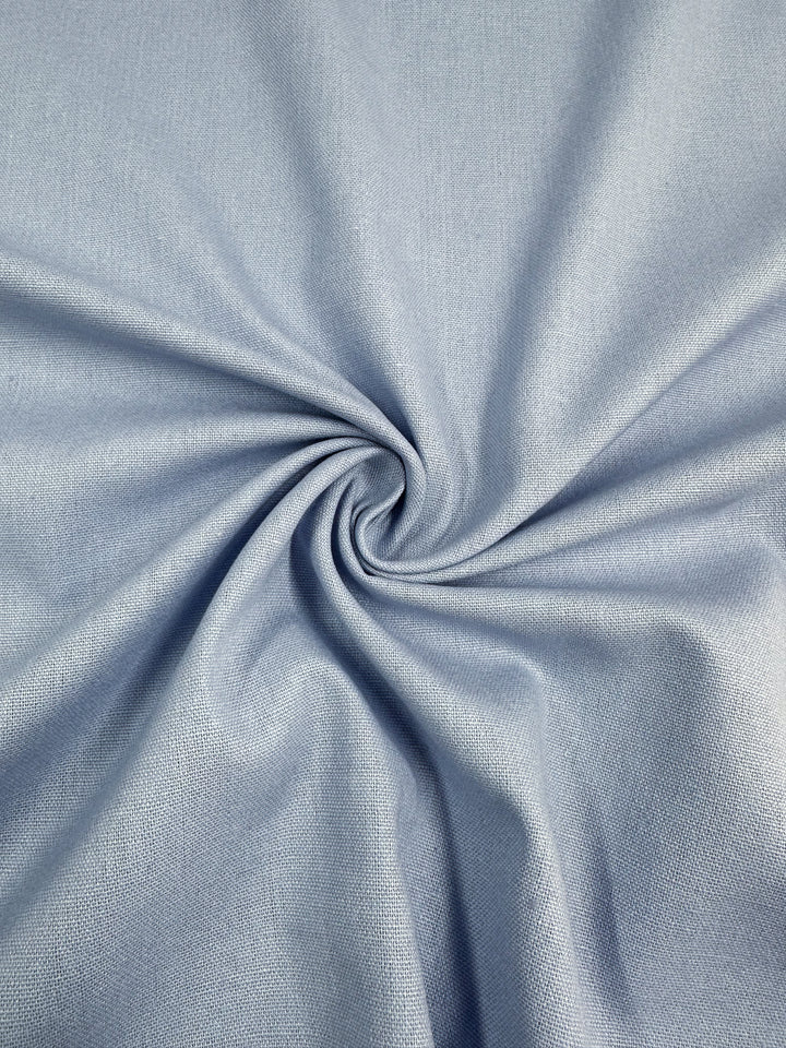 Close-up of the Linen Blend - Skyway from Super Cheap Fabrics, a light blue, medium weight linen fabric with a smooth texture, artistically gathered in a spiral pattern at the center. The folds create a gentle, flowing appearance, highlighting the fabric's soft and slightly shiny surface.