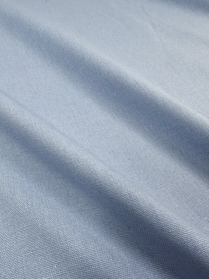 A close-up of Super Cheap Fabrics' Linen Blend - Skyway - 145cm in light blue with soft folds, showcasing the texture and weave of the material. The Italian linen blend fabric appears smooth with subtle, gentle shadows created by the undulations.