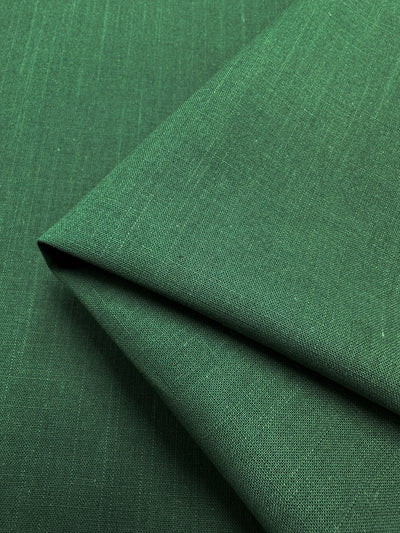 A neatly folded piece of Linen Blend - Fir - 145cm from Super Cheap Fabrics, with a smooth, slightly textured surface, is positioned on a matching green background. The fabric's weave and texture are clearly visible, showcasing its lightweight nature and sharp, crisp edges where it is folded.