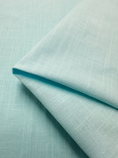 A close-up view of the Linen Blend - Watersprout - 145cm fabric from Super Cheap Fabrics reveals a light blue hue with a subtle textured pattern. The material is neatly folded, showing slightly visible threads and a smooth surface, highlighting its quality craftsmanship made in Italy.