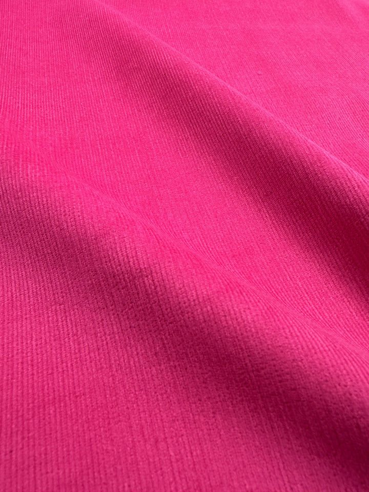 Close-up of the Micro Wale Corduroy - Cabaret fabric by Super Cheap Fabrics, showcasing its ribbed texture. The pink material appears soft and stretches uniformly, highlighting its smooth and subtly ridged surface. Perfect for winter clothing or home decor, the lighting accentuates the subtle shadows and contours, emphasizing its quality at a width of 145cm.