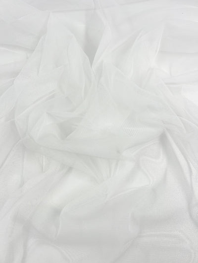 A close-up of sheer white, translucent fabric with delicate, flowing folds. The light weight polyester material appears soft and airy, creating a sense of lightness and elegance. The image highlights the texture and gentle draping nature of the Bridal Tulle - White - 160cm by Super Cheap Fabrics.