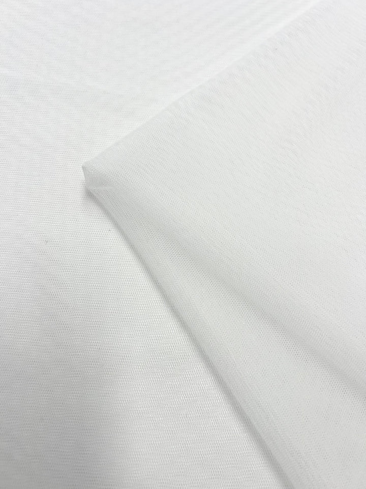 Close-up of sheer white mesh fabric, displaying its fine, see-through texture and grid-like pattern. The Bridal Tulle - White - 160cm from Super Cheap Fabrics appears delicate, with a smooth surface. The edges of the polyester material are slightly folded over the main body.