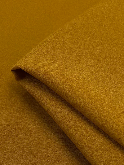 A close-up image of a folded piece of golden brown fabric, likely a Wool Cashmere Blend. The material appears smooth with a subtle texture, giving it a slightly reflective quality. The folds create gentle shadows, adding depth and dimension to the Wool Cashmere - Desert Sun - 150cm by Super Cheap Fabrics, which is dry clean only.