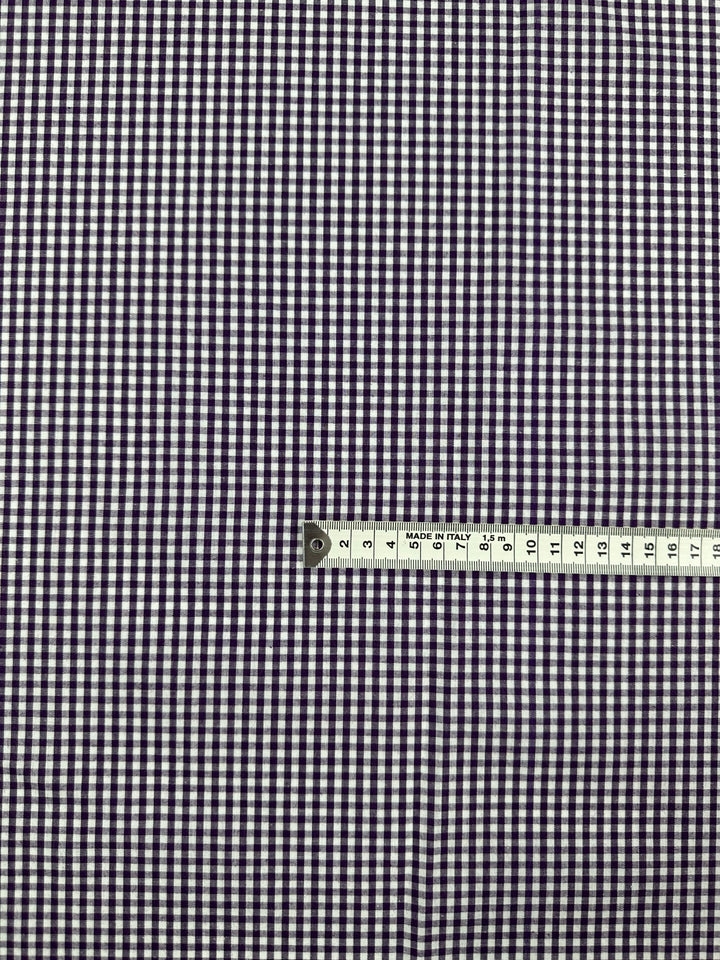 A close-up image of a small checkered gingham fabric pattern in purple and white. A metal measuring tape is placed horizontally across the lower part of this Cotton Gingham - Mini Purple - 140cm, indicating measurements in centimeters. Brand Name: Super Cheap Fabrics