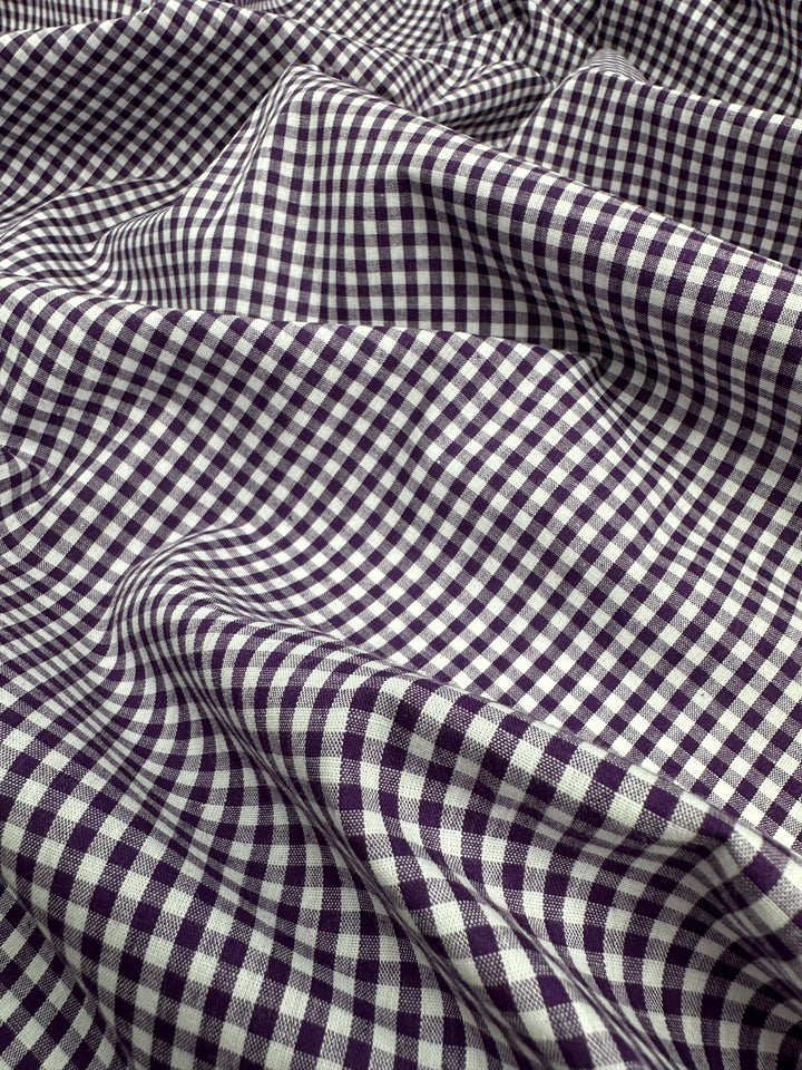 A close-up image of Super Cheap Fabrics' Cotton Gingham - Mini Purple - 140cm with a small checkered pattern in purple and white colors. The 100% cotton fabric appears to be draped and slightly wrinkled, creating a wave-like texture throughout the image.