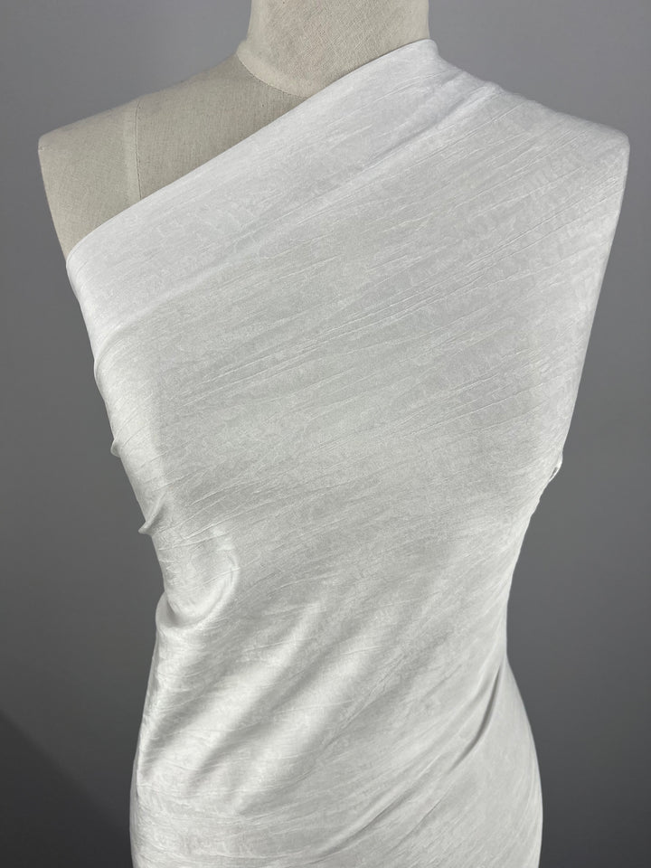 A mannequin adorned in a white dress made from Super Cheap Fabrics' Shimmer Knit material, measuring 150cm and crafted from lightweight polyester spandex, stands elegantly against a gray background. The smooth yet slightly textured fabric enhances the dress's sophisticated allure.