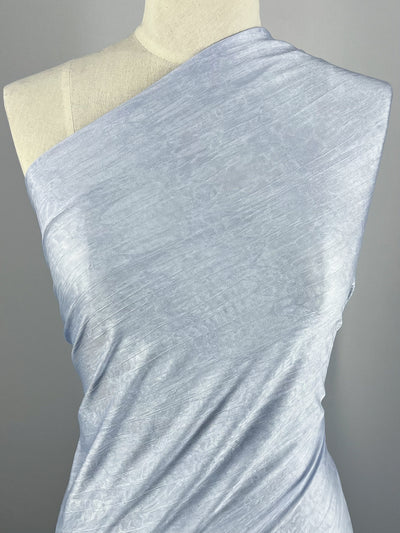 A mannequin is elegantly draped with the Shimmer Knit - Spa - 150cm from Super Cheap Fabrics, a light blue, textured polyester fabric that boasts a subtle sheen. The lightweight fabric is wrapped asymmetrically, covering one shoulder and leaving the other exposed. The plain and neutral background enhances the fabric's vibrant color and intricate texture.