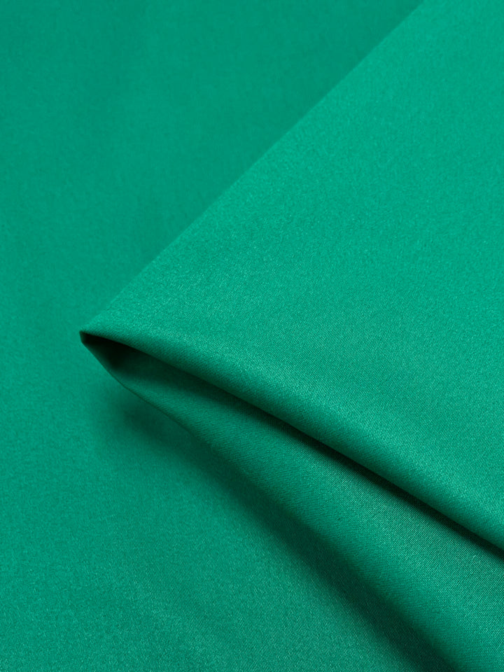 A close-up image of a piece of smooth, green fabric from Super Cheap Fabrics' Plain Sateen collection in "Green Tambourine". The neatly folded material has a subtle sheen that suggests a slightly reflective surface. This 145cm-wide spandex sateen appears to be lightweight and finely woven.