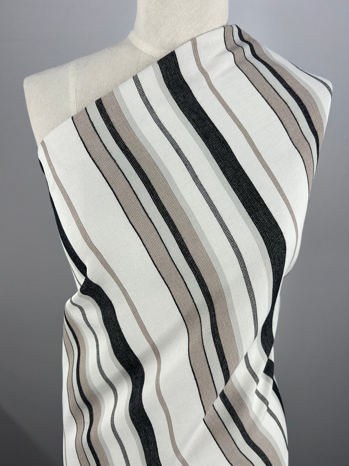 A mannequin is draped in fabric featuring vertical stripes in shades of beige, black, and white. Made from sustainable deadstock materials, the smooth-textured Designer Cotton - Grey Rock Candy - 145cm by Super Cheap Fabrics highlights our commitment to eco-friendly fashion and is displayed against a plain gray background.