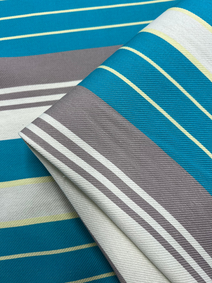 A close-up image of fabric from the Super Cheap Fabrics Designer Cotton - St.Tropez - 157cm Range featuring a pattern of horizontal stripes. The stripes vary in thickness and color, including shades of teal, gray, white, and yellow. The fabric is folded, showing layers of the pattern made from sustainable deadstock materials.