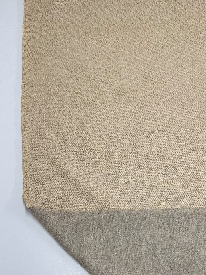 A close-up image of the Unbrushed Fleece - Ancient Scroll by Super Cheap Fabrics, showcasing its beige, ultra-soft texture. The bottom corner of this high-quality 163cm fabric is turned up to reveal the light gray underside. The luxurious fabric is elegantly placed on a smooth, white surface.