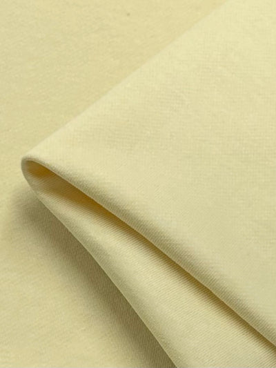 A close-up of a folded piece of light yellow, high-quality cotton fabric from Super Cheap Fabrics' Unbrushed Fleece collection in the French Vanilla color, measuring 160cm wide. The texture appears smooth and soft, with visible stitching along one edge, suggesting it is a finely woven material. The fabric is laid on a flat surface with minimal shadows, highlighting its gentle folds and color.