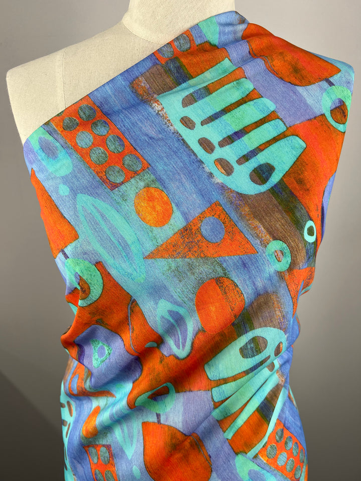 A beige mannequin is draped with a colorful, abstract-patterned fabric, perfect for children's clothing. The light weight fabric features geometric shapes in vibrant shades of orange, teal, and blue, creating a lively, artistic design. The background is plain and neutral, highlighting the Designer Cotton - Fossil - 150cm by Super Cheap Fabrics boldness.