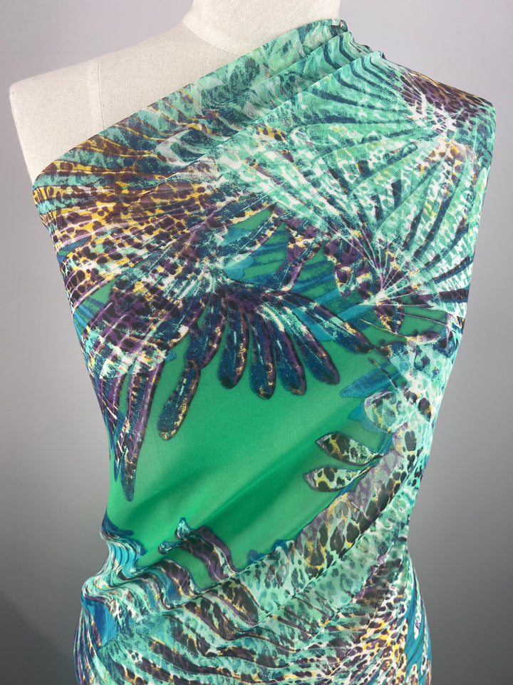 A mannequin is draped in a vibrant, extra-light weight fabric featuring an abstract pattern of green, turquoise, purple, and gold. The design includes feather-like shapes and leopard spots, creating a dynamic and colorful look. This Designer Chiffon - Leopard Palms - 150cm by Super Cheap Fabrics adds to the charm against the neutral gray background.