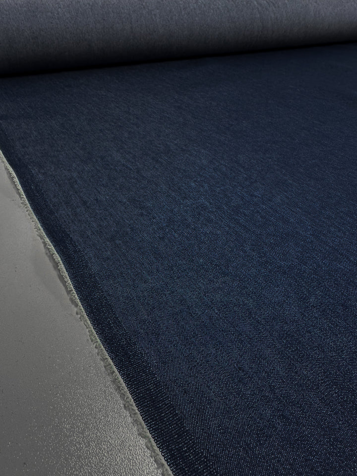 A large roll of Insignia Blue Lightweight Denim from Super Cheap Fabrics, 150 cm wide, is unrolled across a flat surface. The fabric features a raw edge on one side that contrasts with its smooth, uniform texture. Despite its lightweight nature, the material appears thick and durable, making it ideal for the production of jeans.