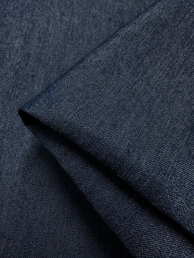 A close-up of Insignia Blue Lightweight Denim from Super Cheap Fabrics showcases the 100% cotton material's fine, diagonal weaving. The fabric is folded, creating shadows and highlights that emphasize its texture and thickness.