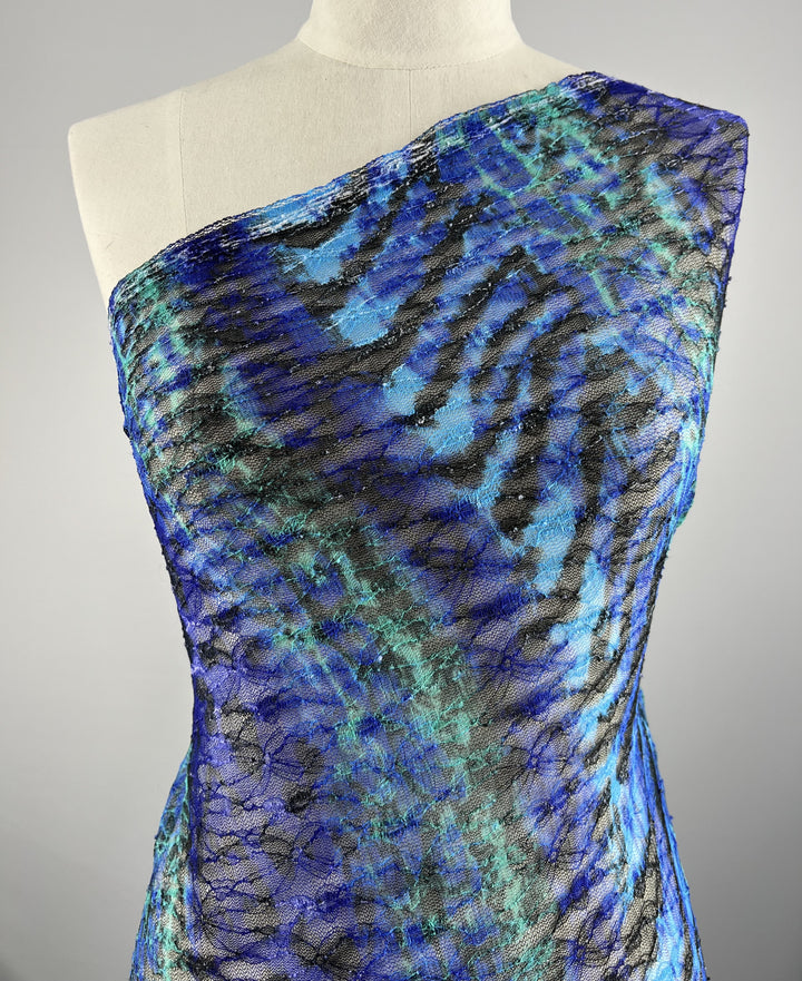 A mannequin showcases the Evening Lace - Avatar - 140cm by Super Cheap Fabrics, featuring a vivid pattern of blue, green, and black abstract stripes. The polyester fabric appears textured, alternating between shimmery and matte sections, creating a dynamic visual effect. The background is plain gray.