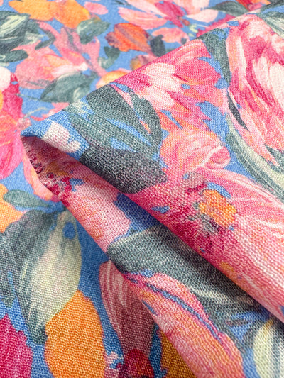 A close-up view showcases the vibrant design of the Super Cheap Fabrics' Soft Cotton - Acrylic - 140cm fabric, featuring a floral pattern in shades of pink, orange, green, and blue. Made from 100% cotton, the slightly folded material highlights its softness and vivid summer design. The background also displays more of this captivating floral theme.