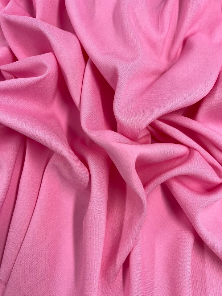 Image shows a close-up of Super Cheap Fabrics' Plain Rayon in Prism Pink, 145cm wide, displaying its soft folds and rich texture. The lighting accentuates the smooth surface and the varying shades of pink, creating a sense of depth and fluidity characteristic of lightweight fabrics.