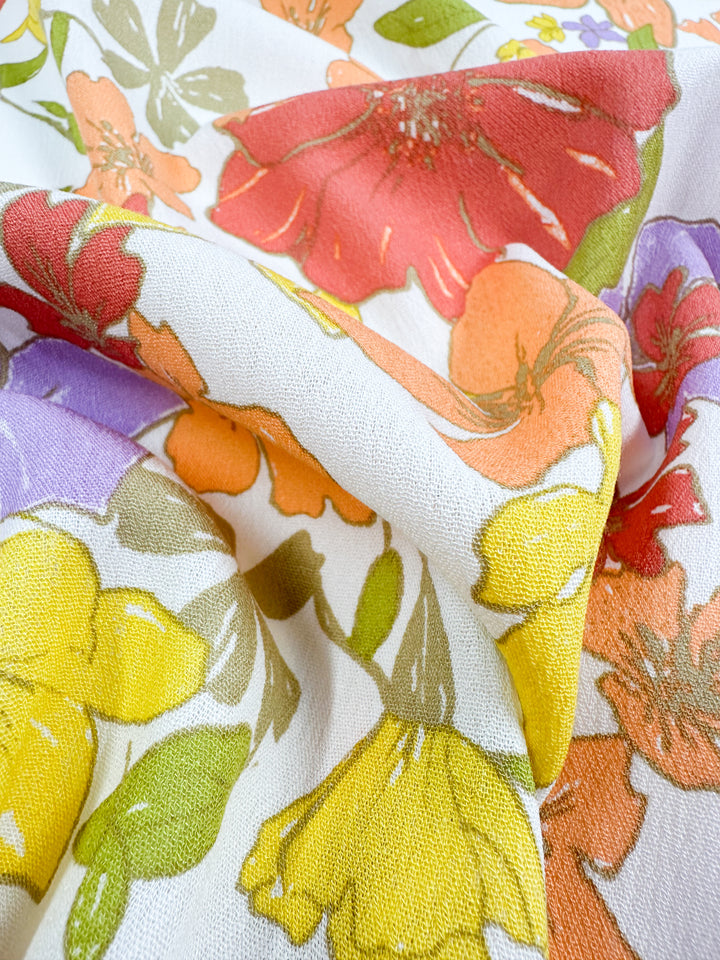 A close-up of Soft Cotton - Illustrator - 140cm by Super Cheap Fabrics reveals a colorful floral masterpiece. Crafted from soft cotton, the fabric showcases a pattern of large, vibrant flowers in shades of orange, red, yellow, and purple, accompanied by green leaves on a pristine white background. Slightly wrinkled for added texture, this breathable summer fabric is as comfortable as it is visually stunning.