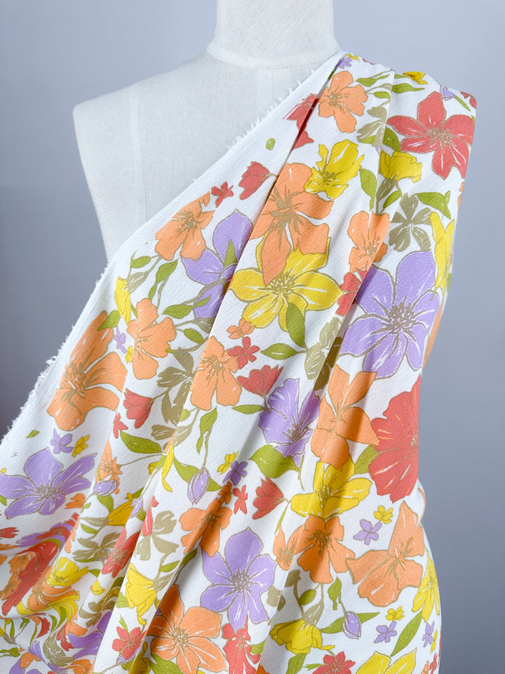 A white mannequin is draped with Super Cheap Fabrics' Soft Cotton - Illustrator - 140cm, adorned with a colorful floral pattern. The flowers are in shades of orange, yellow, pink, red, and purple, creating a vibrant and lively design. The breathable fabric is perfect for summer wear. The background behind the mannequin is plain and light gray.