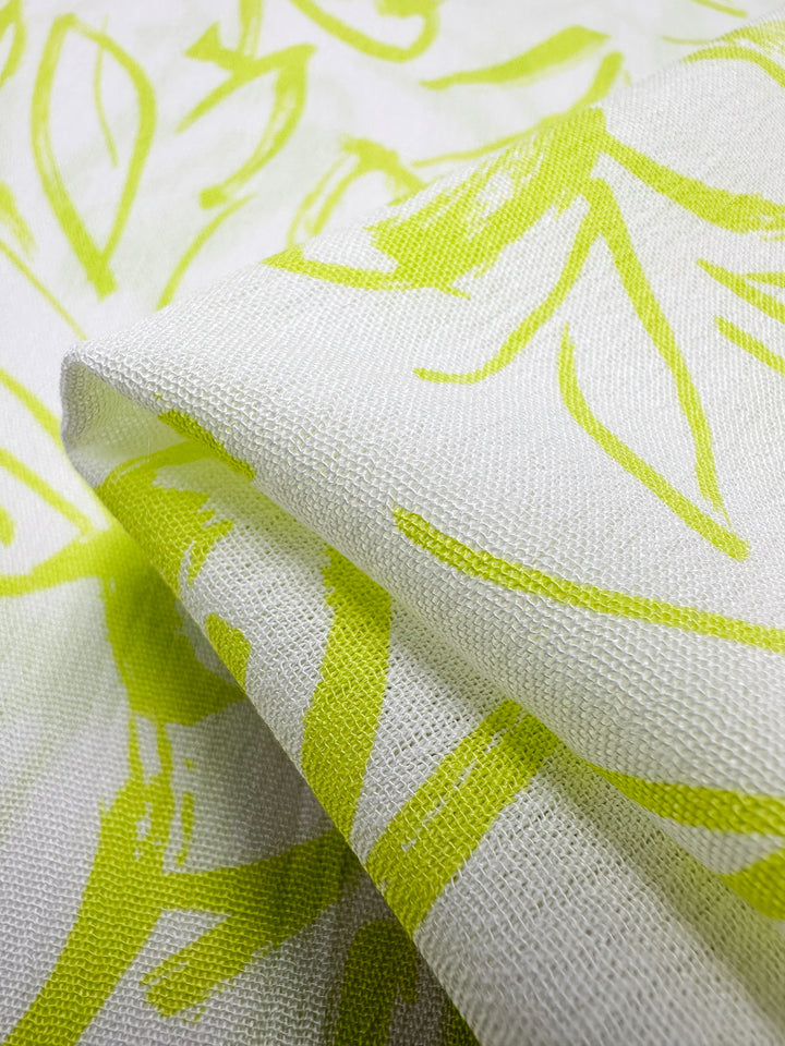 A close-up of a piece of textured fabric with a white base color. The Soft Cotton - Love Bird - 140cm from Super Cheap Fabrics is adorned with abstract yellow-green botanical patterns. It is slightly creased, showcasing its texture and the intricacy of the design print, making it evident that it's made from breathable fabric.