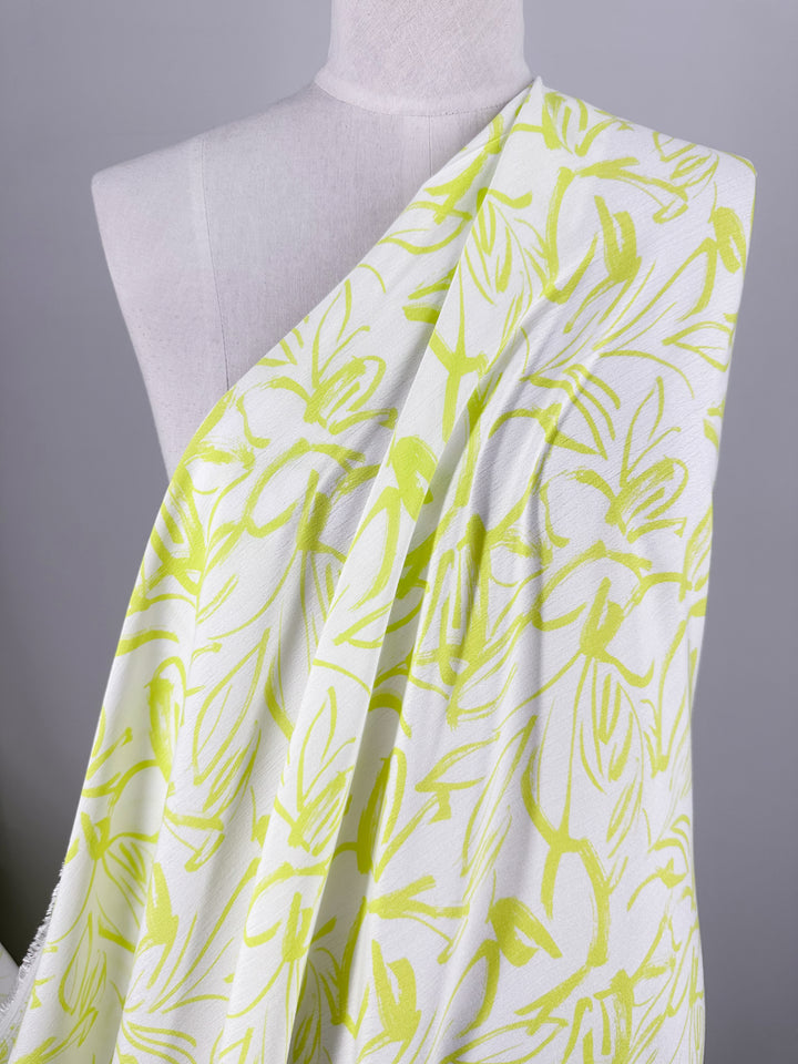 A mannequin is draped in the Soft Cotton - Love Bird - 140cm fabric by Super Cheap Fabrics, showcasing a lively, abstract green pattern on a white 100% cotton background. The design features various overlapping, sketch-like lines and brushstrokes that create a dynamic visual effect, with the plain background highlighting the intricate details of this summer fabric.