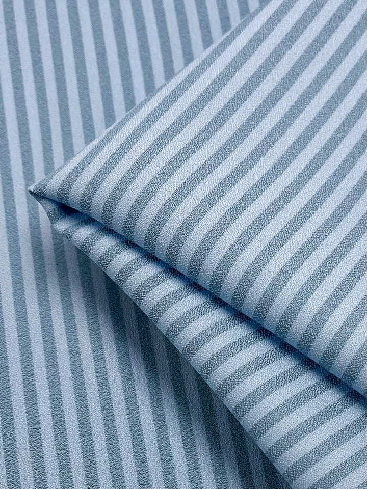 The "Striped Crepe - Milky Blue - 150cm" by Super Cheap Fabrics showcases a neatly folded polyester fabric in a milky blue hue, featuring alternating light and dark blue stripes that form a textured pattern. This smooth and soft fabric exudes high fashion and is an ideal option for clothing or home textiles.