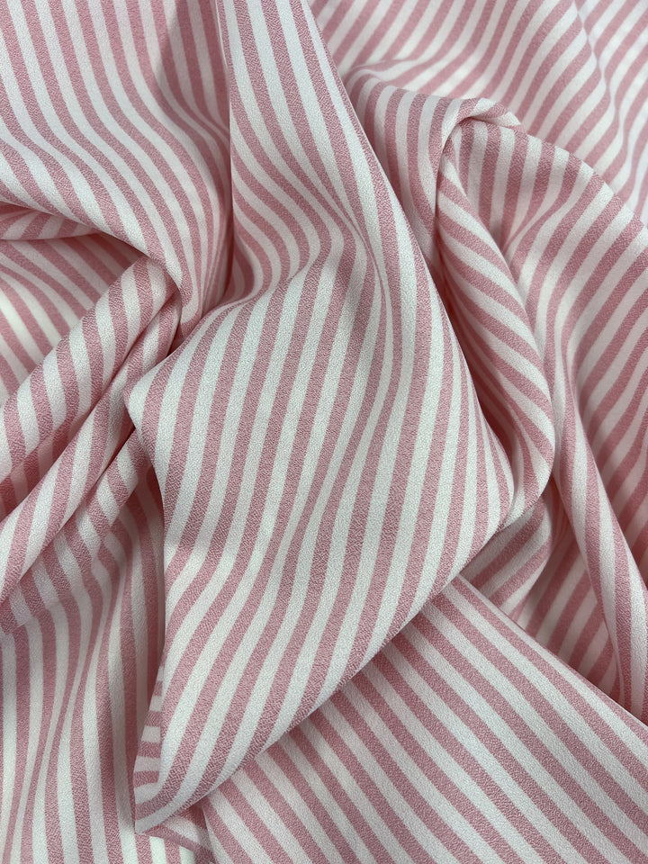 A detailed view of the Striped Crepe - Tickled Pink fabric by Super Cheap Fabrics reveals a soft polyester material with diagonal pink and white stripes, creating a dynamic and fashionable pattern.