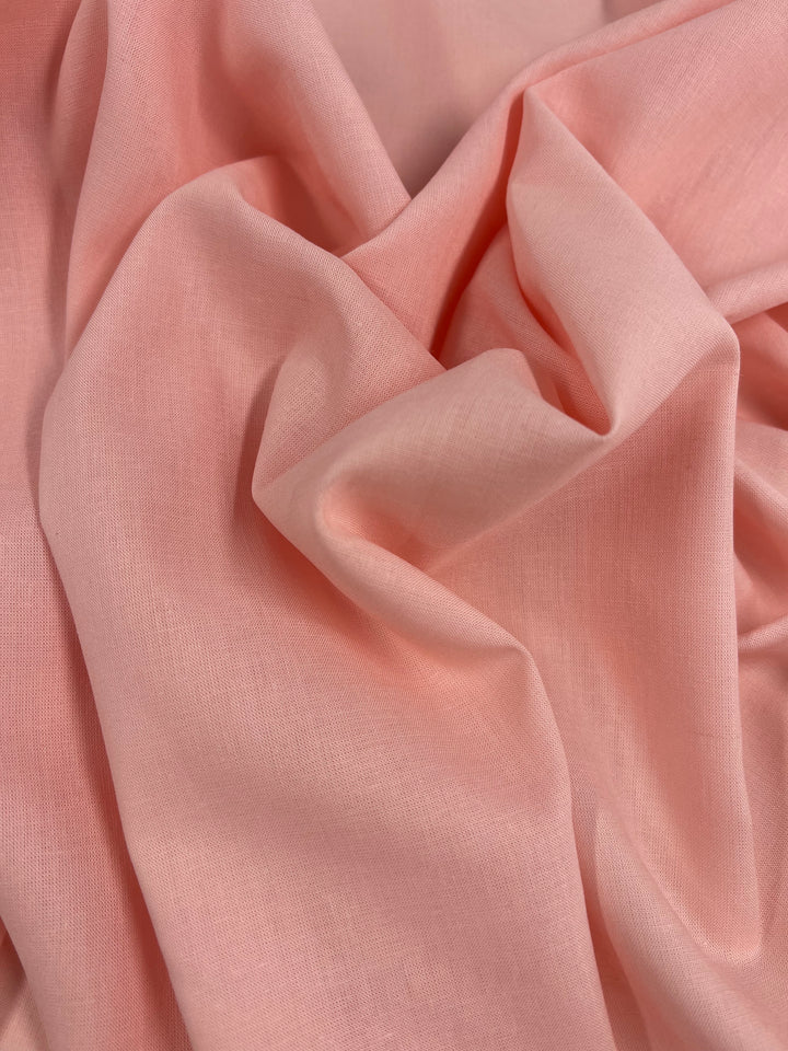 A close-up photograph showcases the Pure Linen - Salmon Pink fabric from Super Cheap Fabrics. The material displays gentle folds and a smooth texture. Made from 100% linen, it appears lightweight and semi-sheer with a slightly crinkled look, measuring 140cm wide.