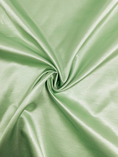 A close-up image of the Shantung Satin - Foam Green - 150cm by Super Cheap Fabrics. The lightweight material is smooth and shiny, with soft folds forming a circular pattern in the center, creating an elegant and flowing appearance. The lighting accentuates the fabric’s sheen and texture.