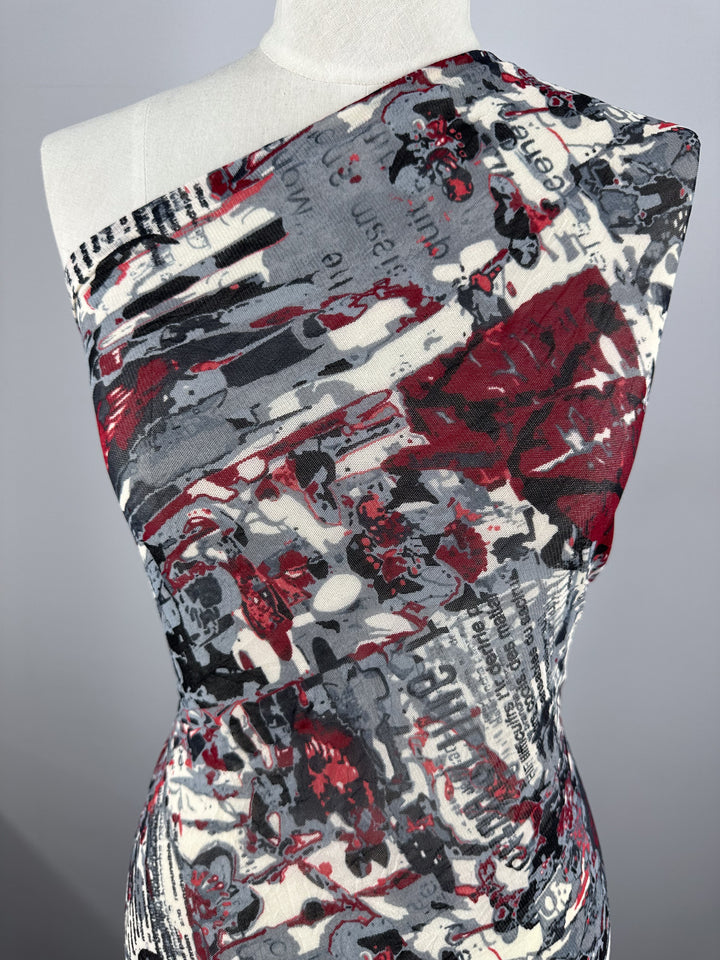 A mannequin is draped in Super Cheap Fabrics' Printed Mesh - Read - 120cm, a lightweight fabric showcasing a bold, abstract print in red, black, and white. The sheer material is asymmetrically wrapped to cover one shoulder, revealing intricate and chaotic patterns reminiscent of newspaper text interspersed with artistic paint splatters.