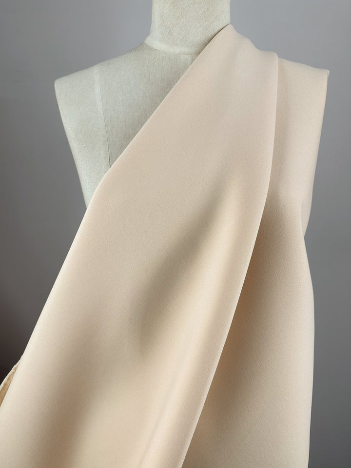 A close-up view of Super Cheap Fabrics' Scuba Knit - Mother of Pearl - 165cm draped elegantly over a mannequin. The smooth, heavyweight fabric falls in soft folds, highlighting its texture and subtle sheen against the minimalist gray background.