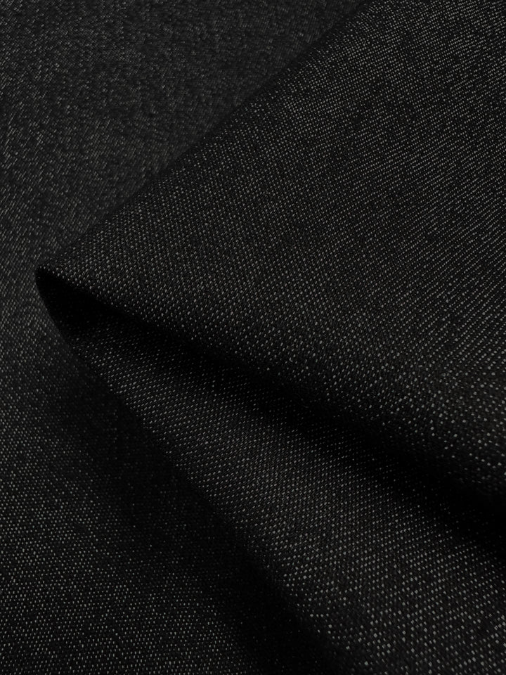 Close-up of a piece of black, woven fabric with a subtle sheen. The fabric, named Lightweight Denim - Black - 135cm by Super Cheap Fabrics, is made from 100% cotton and is folded to highlight its texture and thickness. The weave pattern adds depth to the lightweight denim fabric, while the lighting accentuates its smooth, matte finish.