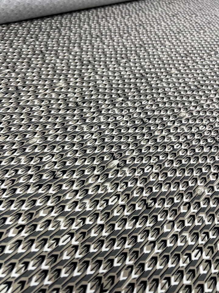 A close-up of a textured surface featuring a repeating, intricate pattern of interlocking diamond shapes. The colors are primarily silver and gray, creating a metallic appearance. This medium-weight fabric, Super Cheap Fabrics' Stretch Bengaline - Directions - 150cm, has a slightly reflective sheen that gives it a glimmering effect.