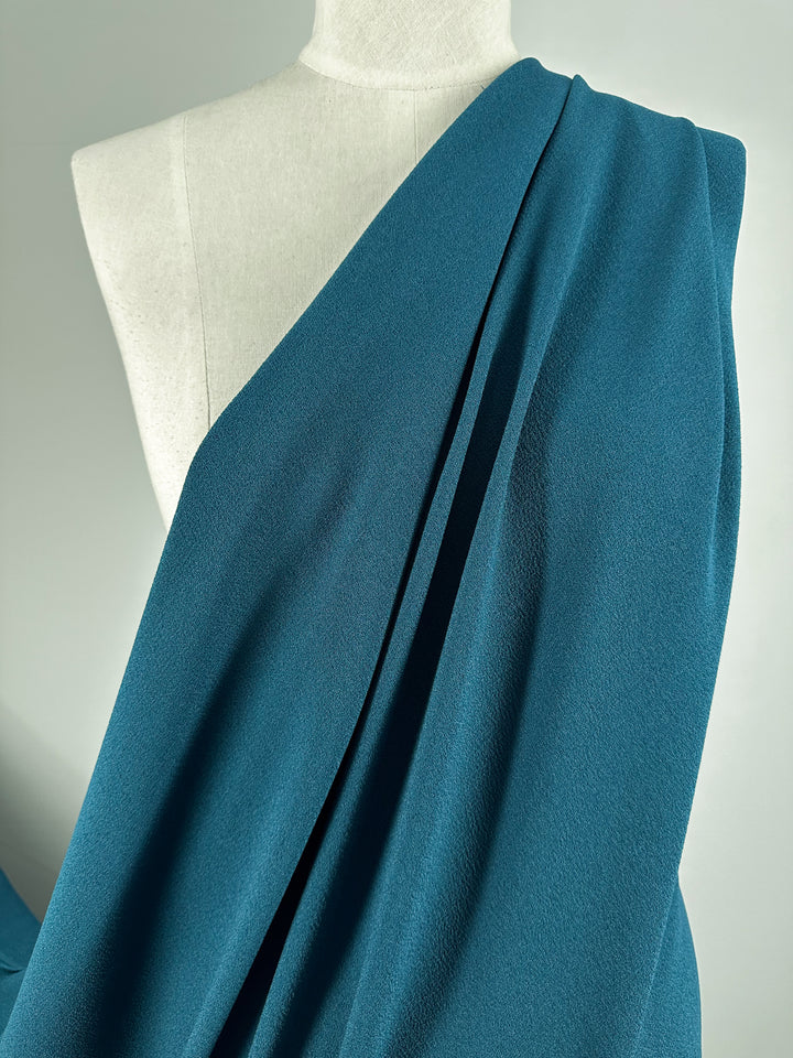 A minimalist torso mannequin draped with Designer Crepe - Teal - 145cm from Super Cheap Fabrics, showcasing its soft, flowing polyester texture. The elegant fabric is arranged over one shoulder, creating a simple yet refined visual display against a pale gray background.