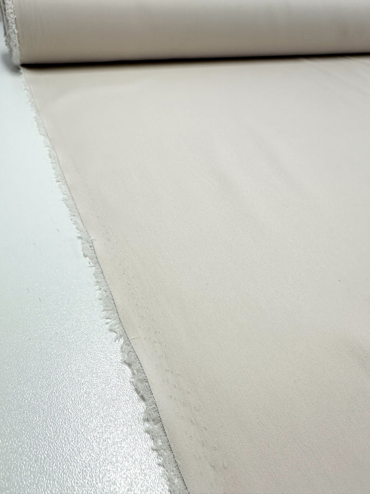 A roll of Stretch Cotton - Perfectly Pale from Super Cheap Fabrics is partially unrolled on a flat surface. The edge of the light beige, medium to heavy weight knit fabric on the left is slightly frayed, and the texture appears smooth and even. The color is consistent across the entire visible portion, making it perfect for crafting crisp fitted dresses.