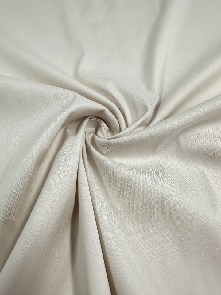 A close-up of "Stretch Cotton - Perfectly Pale - 137cm" from Super Cheap Fabrics shows the pale, light beige medium to heavy weight knit fabric twisted into a spiral. The material appears smooth and slightly shiny, with subtle folds emanating from the center of the twist.