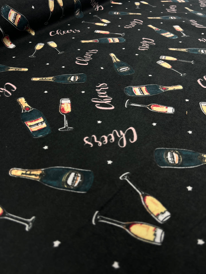 A close-up of lightweight black fabric with a festive pattern featuring champagne bottles, champagne glasses filled with bubbly, and the word "Cheers" written in cursive. The 100% cotton design also includes small white star accents scattered throughout. This is the Flannelette - Cheers - 110cm by Super Cheap Fabrics.