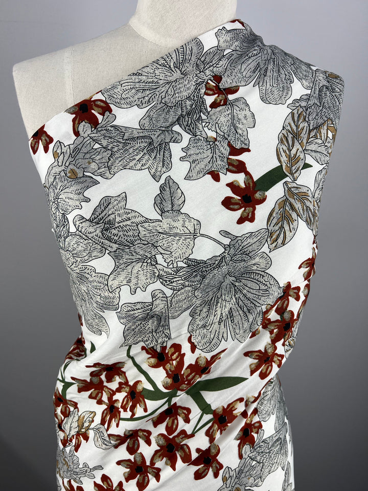 A draped mannequin showcases the versatile elegance of "Printed Rayon - XL Flora - 145cm" from Super Cheap Fabrics, adorned with an intricate pattern featuring large, detailed leaves in shades of gray and beige, interspersed with clusters of red flowers and green stems on a white background.