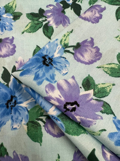 A close-up shot of the Super Cheap Fabrics Printed Rayon - Watercolour (145cm), showcasing a versatile light blue fabric adorned with vibrant prints of blue and purple flowers interspersed with green leaves. A folded section reveals the underside, which features the same stunning floral pattern.