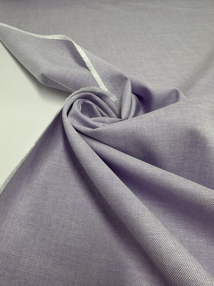 Super Cheap Fabrics’ Oxford Cotton in Baby Lavender (146cm) is shown spread out with soft folds and a gentle swirl, highlighting its subtle texture. The light purple fabric displays a visible white selvage along the top left.