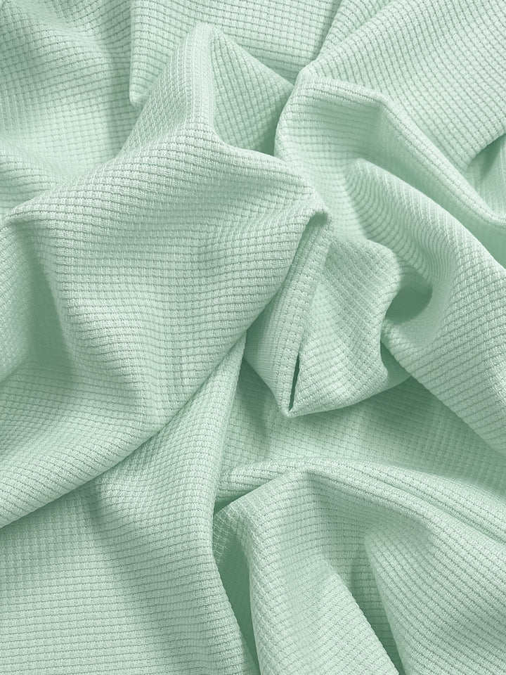 A close-up image of light green textured fabric, displaying a crinkled and folded appearance. The fabric appears to have a waffle pattern, creating a visually interesting and three-dimensional appeal. This is the Waffle Knit - Ambrosia - 170cm from Super Cheap Fabrics.