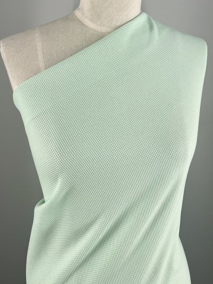 A mannequin is displayed wearing a Waffle Knit - Ambrosia - 170cm by Super Cheap Fabrics, which is a one-shoulder, light mint green top with a three-dimensional appearance. The top drapes stylishly over one shoulder, leaving the other exposed. The background is plain and gray, providing contrast to highlight the textured garment.
